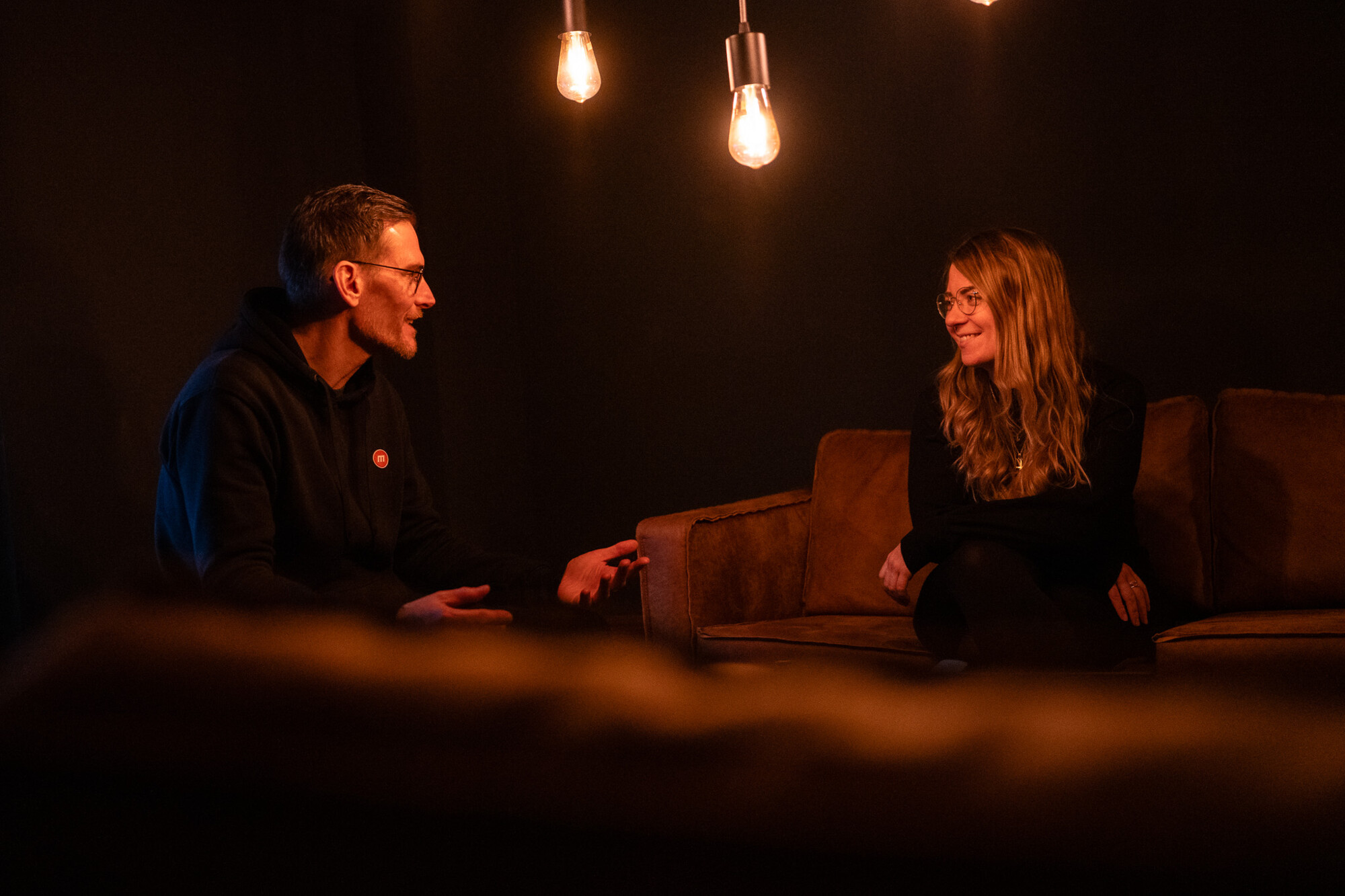Zwei Personen im warm beleuchteten Raum unterh alten sich entspannt auf braunem Sofa vor dunklem Hintergrund.