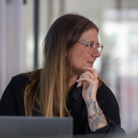 Frau mit Brille und Tätowierungen denkt nach, sitzend vor Laptop in moderner Arbeitsumgebung.