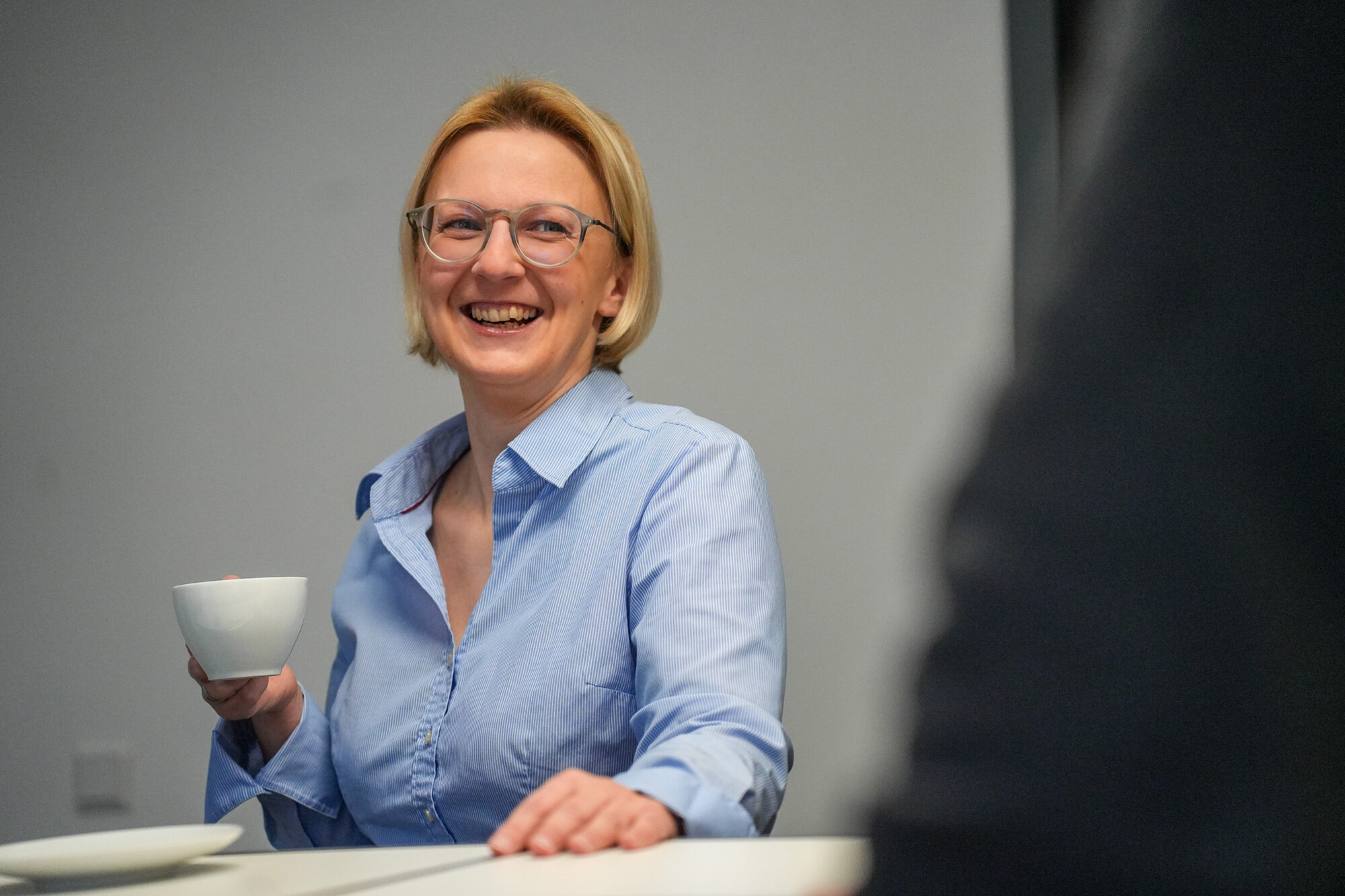 Frau mit blauer Bluse und Brille lacht entspannt und hält eine weiße Tasse Kaffee in der Hand.