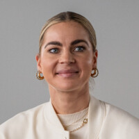 Junge Frau mit blondem Zopf, goldenen Creolen und dezentem Make-up vor neutralem Hintergrund lächelnd.