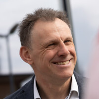 Mann im Anzug lächelt freundlich im Freien vor unscharfem Hintergrund mit Windkraftanlagen.