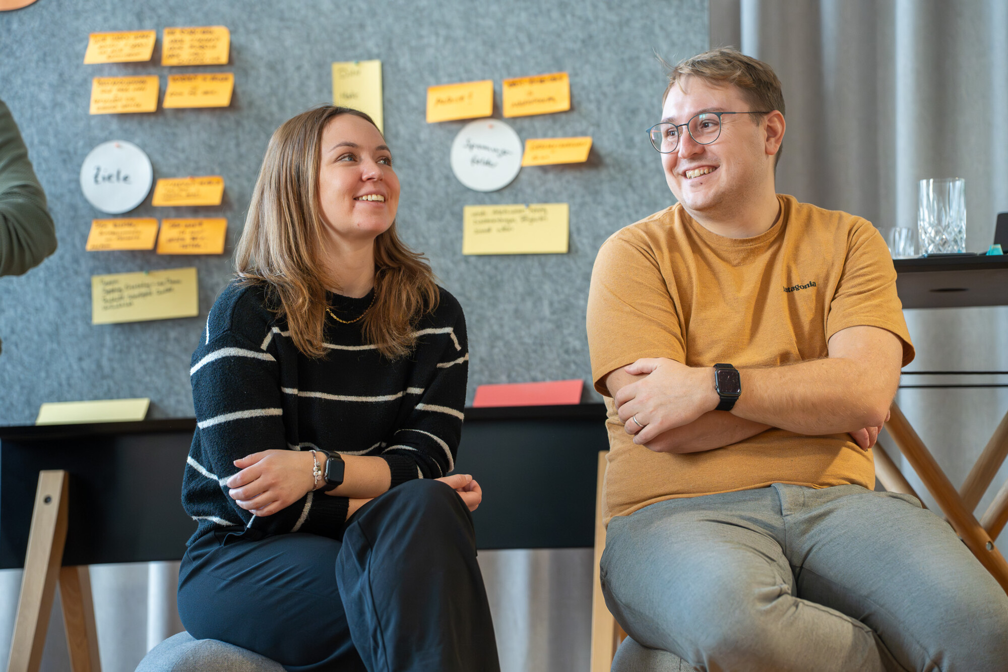 Zwei junge Personen sitzen entspannt im Meeting, lächeln und tauschen Ideen vor einer Wand mit Notizzetteln aus.