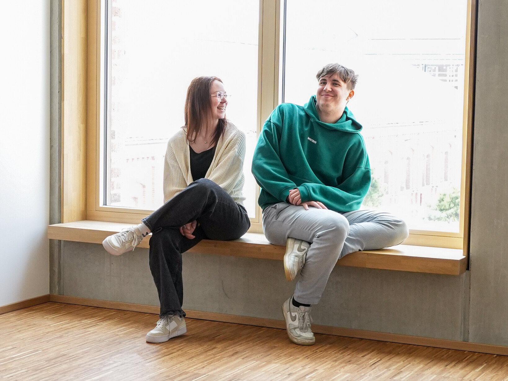 Zwei junge Menschen, lächelnd und entspannt, sitzen auf einer Fensterbank in heller, moderner Umgebung.