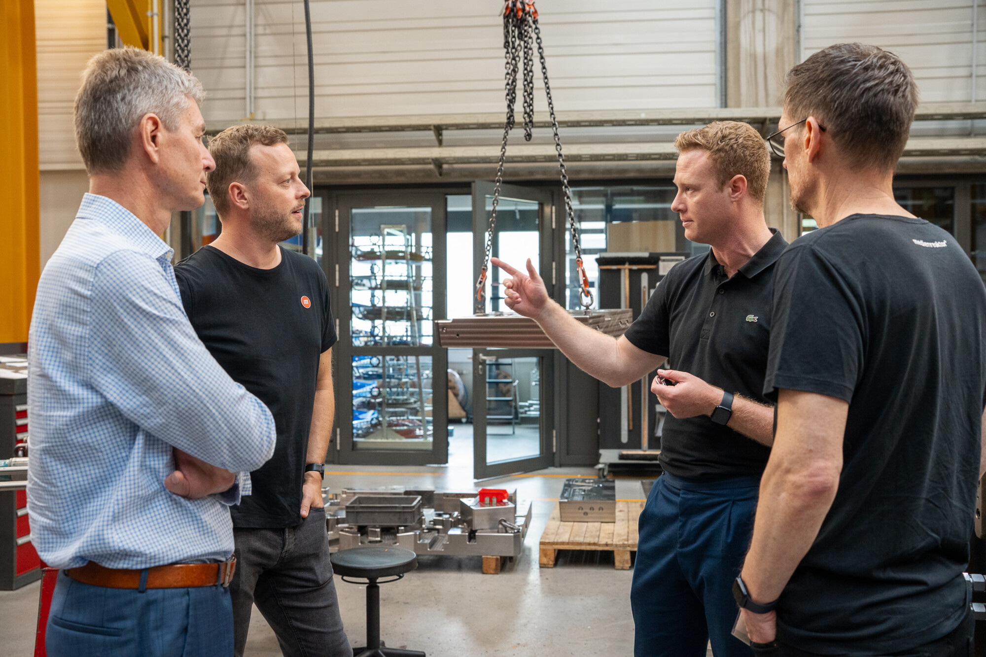 Vier Männer diskutieren in moderner Industriehalle über technische Bauteile, Fokus auf Teamarbeit und Planung.
