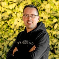 Mann mit Brille und schwarzem Hoodie mit „Deutschlands Dienstrad“-Logo vor grüner Pflanzenwand, freundlich lächelnd.