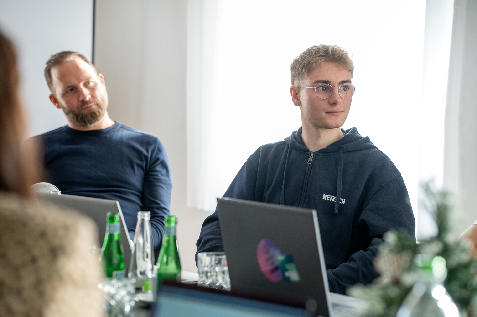 Junge Männer im Meeting mit Laptops und Getränken, konzentriert und aufmerksam in hellem Besprechungsraum.