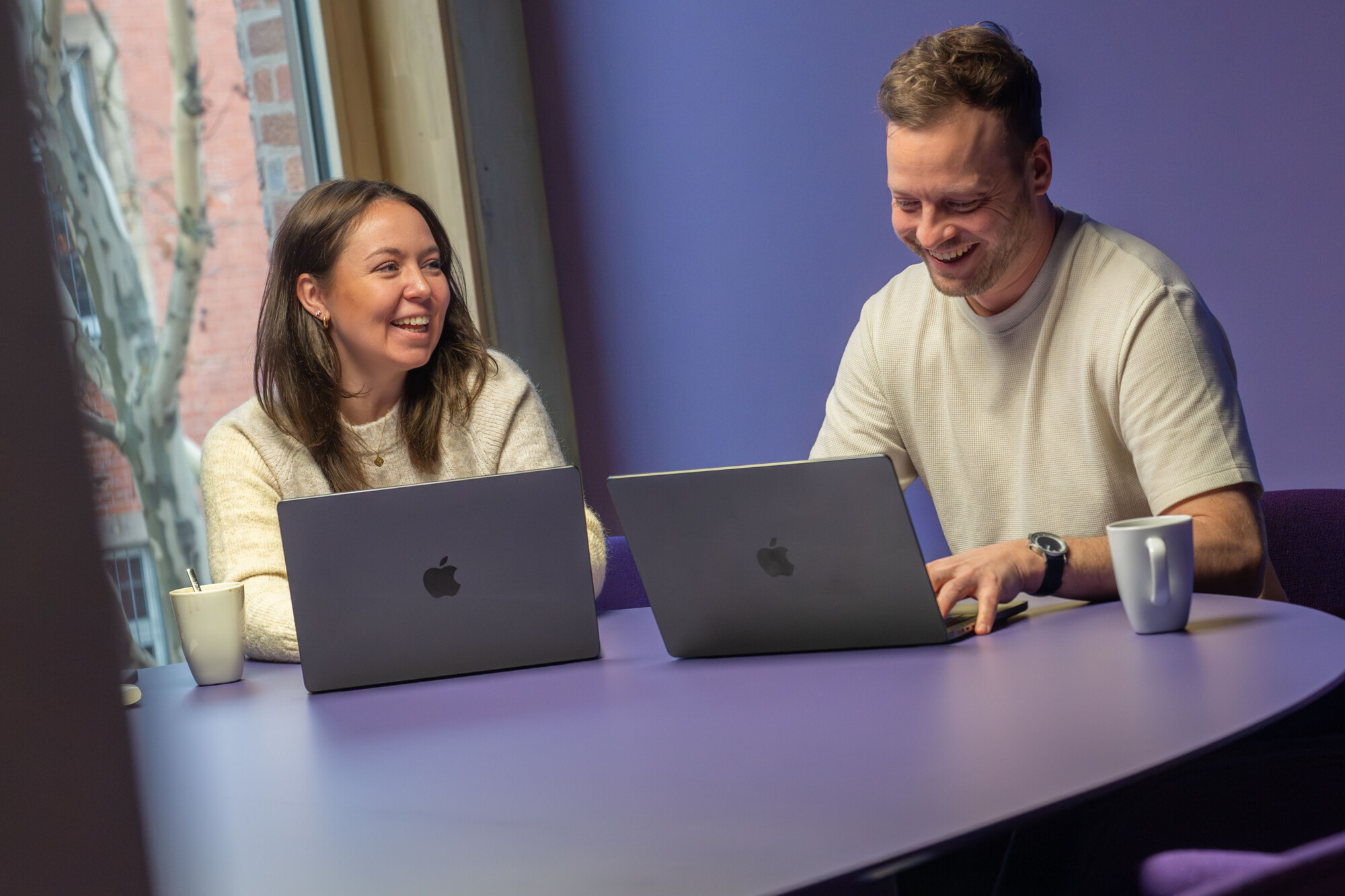 Zwei junge Erwachsene arbeiten lächelnd an ihren Apple MacBooks an einem lila Tisch in heller Büroumgebung.