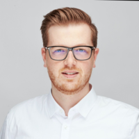 Mann mit rotbraunem Haar und Bart trägt moderne Brille mit Blaulichtfilter, trägt weißes Hemd vor neutralem Hintergrund.