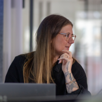 Frau mit Brille und Tattoos denkt aufmerksam bei der Arbeit im modernen Büro mit Laptop und hellem Hintergrund.