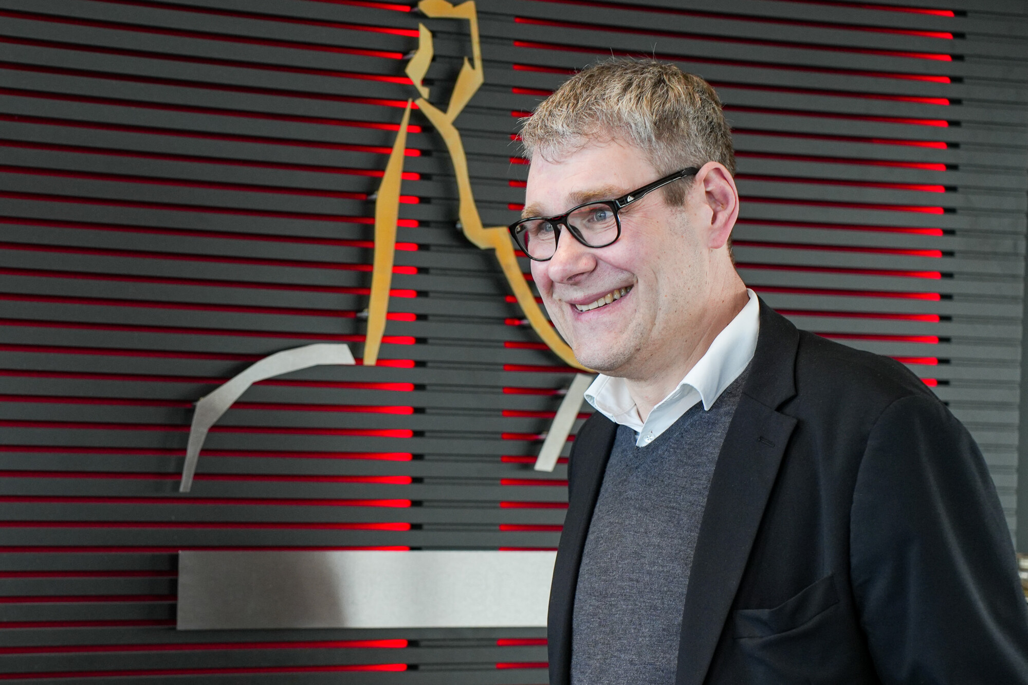 Mann mit Brille und Business-Outfit lächelt vor moderner Wandgestaltung mit roter Beleuchtung und Metallkunst.