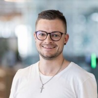 Junger Mann mit Brille und Kreuzkette lächelt freundlich vor unscharfem Bürohintergrund, zukunftsorientiert und sympathisch.